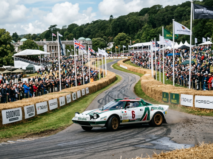 Lancia Stratos rally car number 6 racing on a curving track at the Goodwood Festival with spectators behind hay bales and flags.