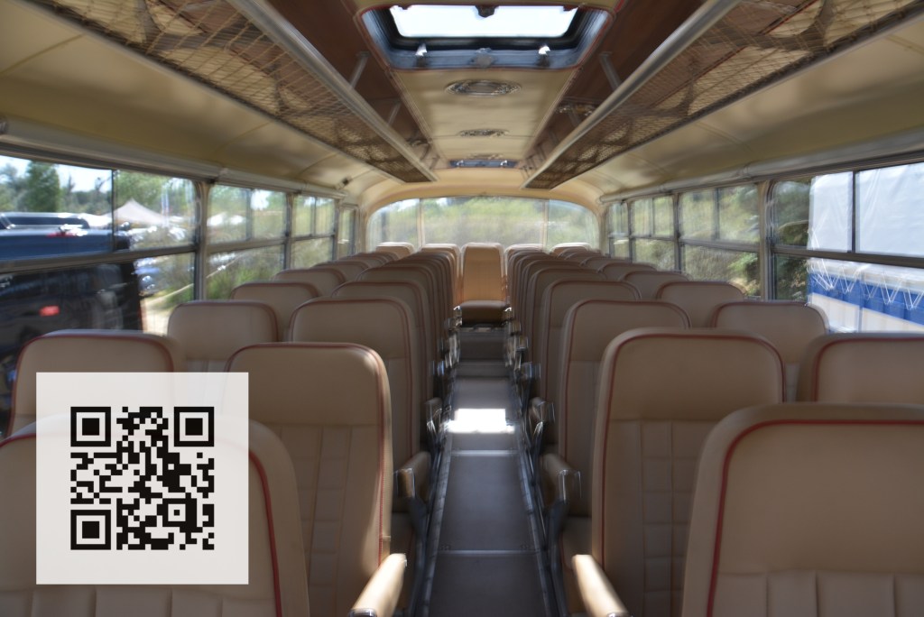 Interior view of a vintage bus showcasing rows of tan upholstered seats, with a clear aisle running down the middle. Natural light is coming through the windows above the seats.