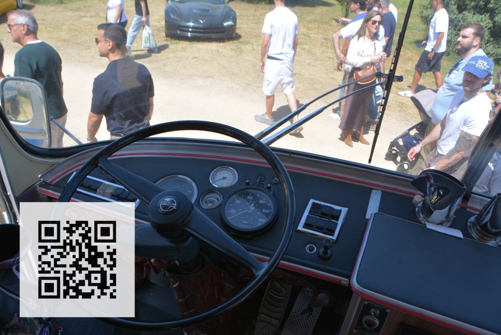 The interior view of a vehicle's cockpit, featuring a steering wheel, gauges, and controls, with a busy outdoor scene visible through the front windshield. A QR code is displayed in the lower left corner.