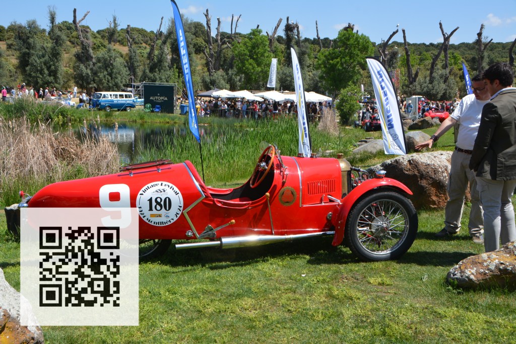 A vintage red car with a number 9 displayed, parked on grass near a pond, while two people are conversing nearby. In the background, there are event tents and spectators enjoying the sunny day.