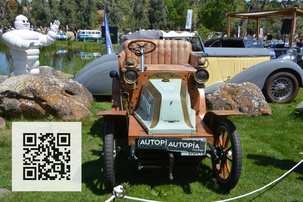 A vintage car displayed at an outdoor event, featuring a wooden body and leather seats, with a Michelin mascot waving in the background, surrounded by greenery and other classic cars.