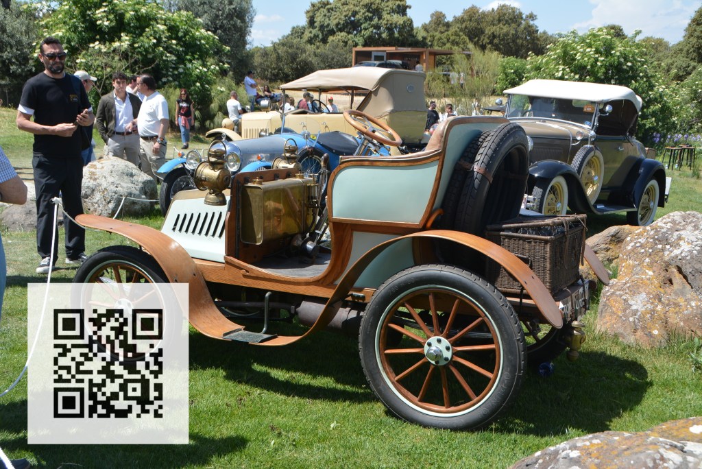 A vintage car displayed on grass, featuring a wooden frame and brass details, with spectators and other classic cars in the background.