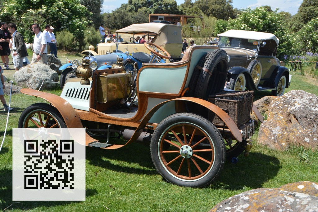 A vintage car with a wooden frame and classic design parked on grass, surrounded by people and other antique vehicles in the background.