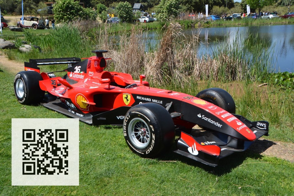 A red Formula 1 racing car displayed on grass near a pond, with tall grass and trees in the background. A QR code is visible in the corner of the image.
