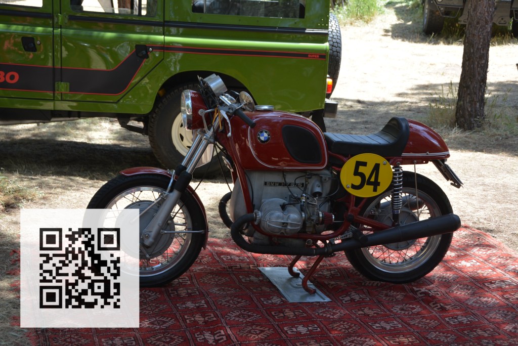 A vintage BMW motorcycle displayed on a patterned rug, featuring a yellow number 54 on its side. In the background, a green vehicle is partially visible.