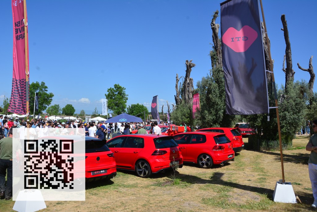 A lively outdoor car event featuring a gathering of people, several red Volkswagen Golf GTI cars, and promotional flags in a sunny park setting.