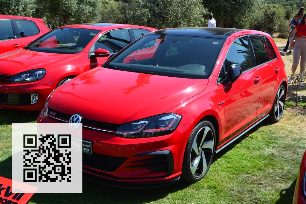 A red Volkswagen Golf GTI parked on green grass, with several other red cars in the background and a QR code visible in the foreground.