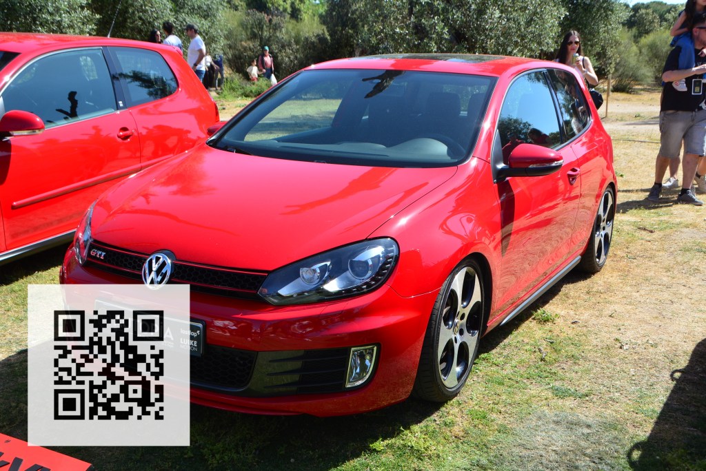 A bright red Volkswagen Golf GTI parked outdoors, featuring sleek design and stylish alloy wheels. In the foreground, a QR code is visible, along with blurred figures of attendees in the background.