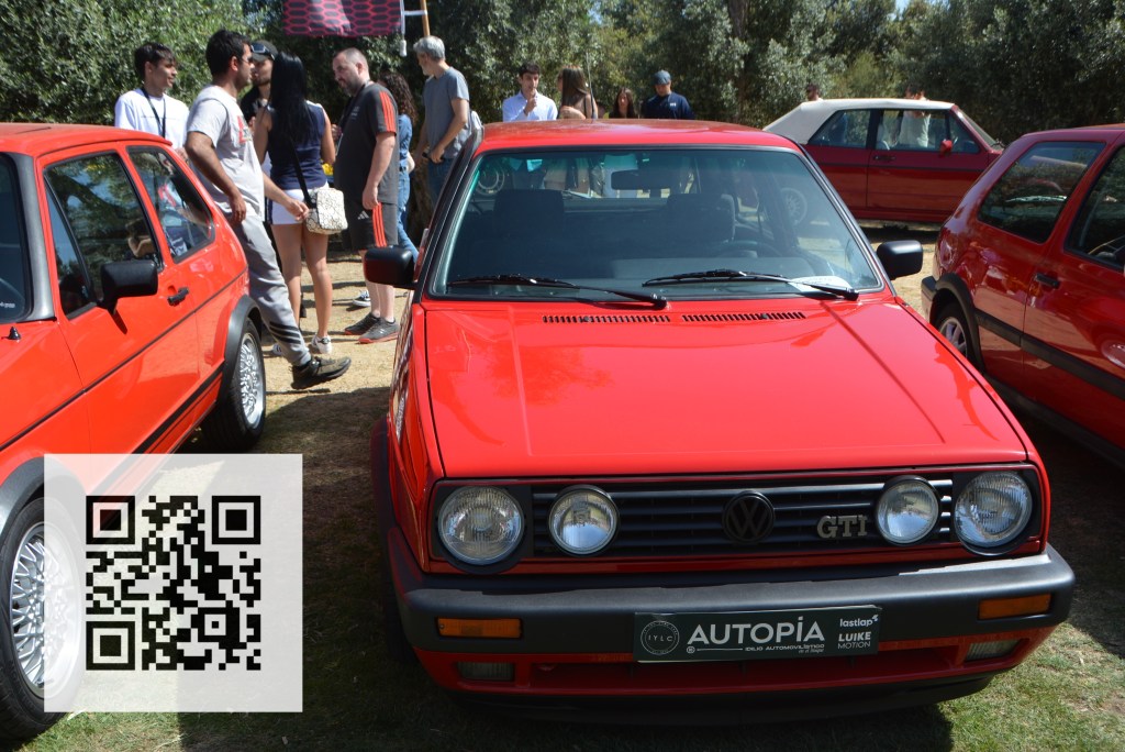 A bright red Volkswagen GTI car parked at an outdoor event, with a QR code in the lower left corner and several people admiring cars in the background.