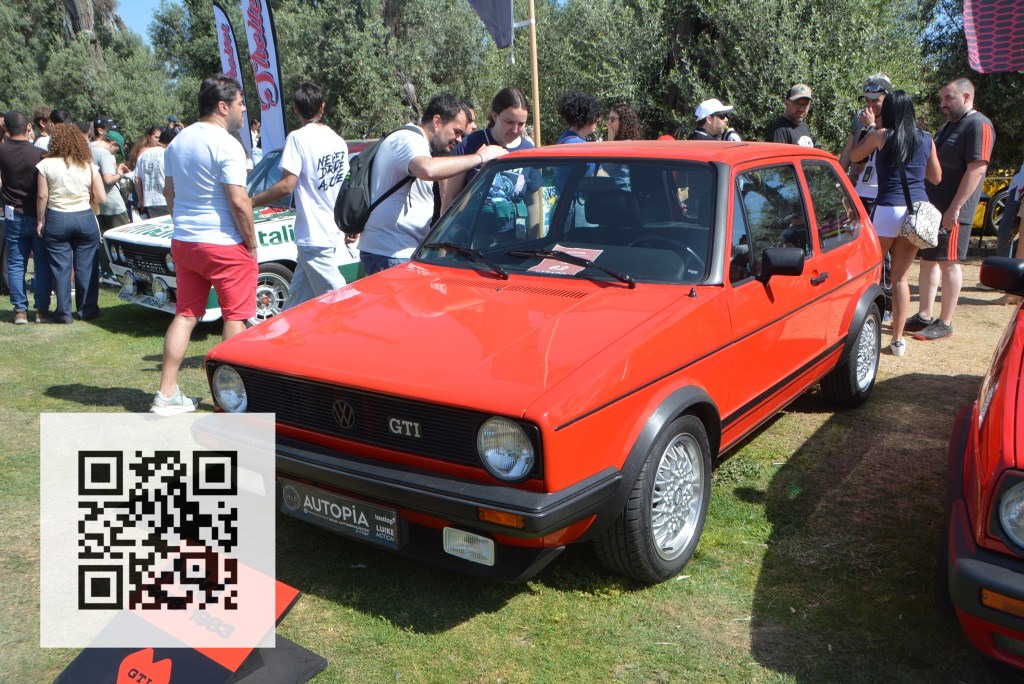 A red Volkswagen GTI parked at an outdoor event, with people admiring it and other cars in the background. A QR code is visible in the bottom corner.