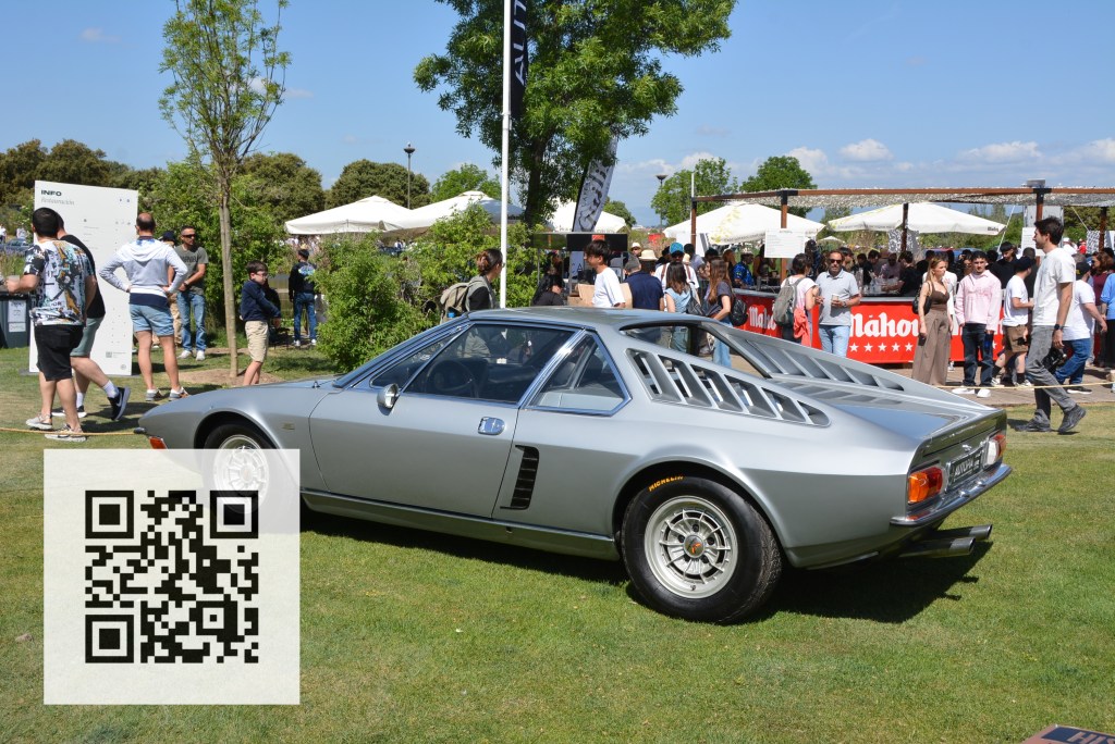 A classic silver sports car displayed on a grassy area, with a group of people in the background enjoying an outdoor event. A QR code is visible on the bottom left corner.