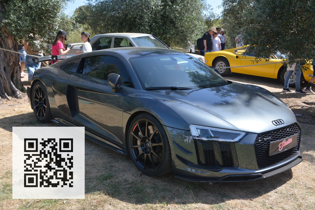 A sleek dark grey Audi R8 sports car parked on grass, with a QR code displayed in the foreground. In the background, people are gathered near other classic cars, including a yellow sports car.