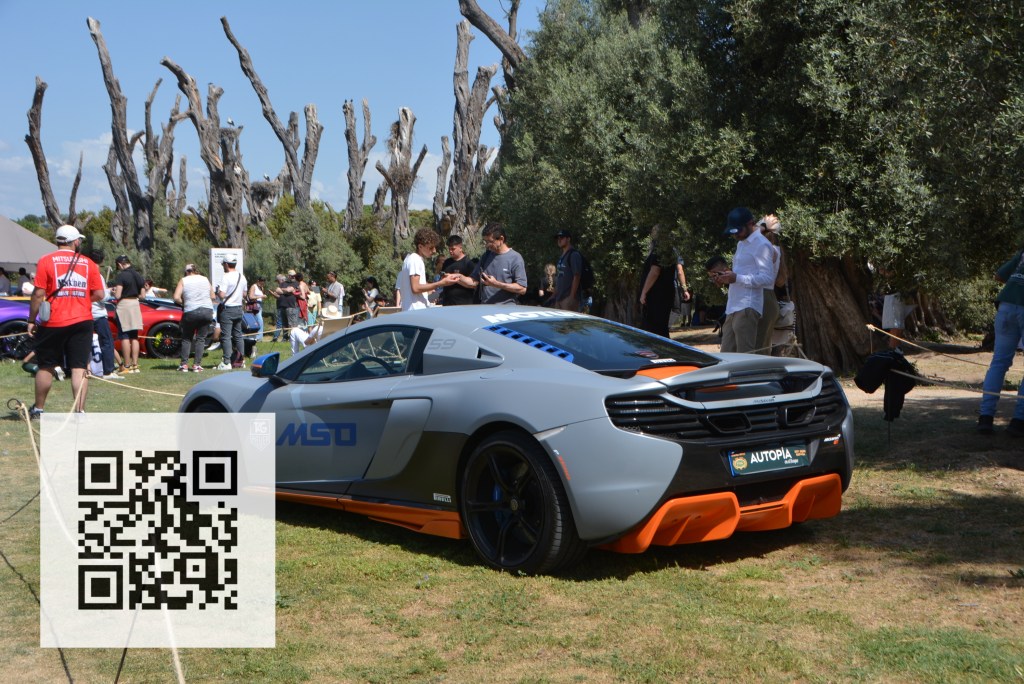 A McLaren sports car with a gray and orange color scheme, showcased at an outdoor event with spectators in the background and olive trees.