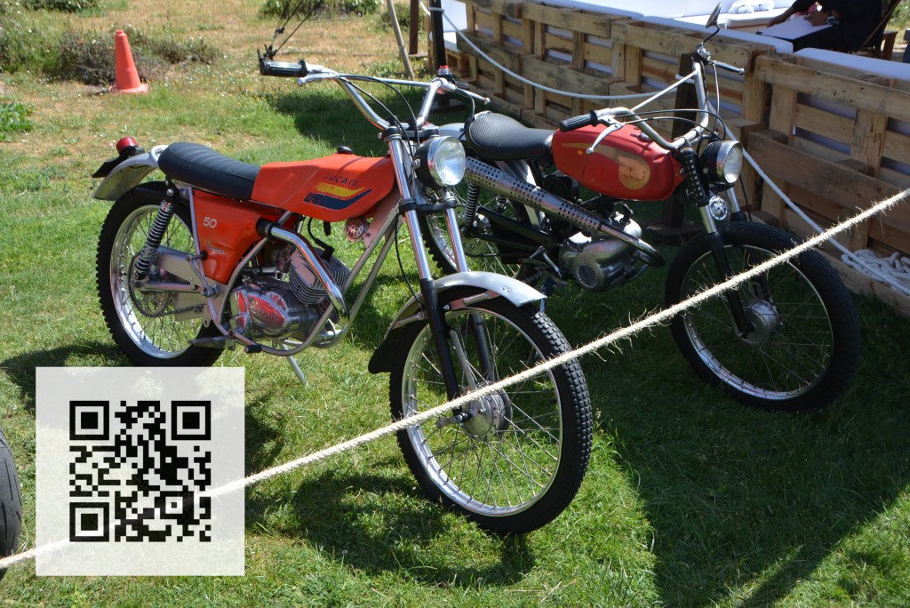 Two vintage motorcycles displayed on grass, featuring an orange Ducati model and a black motorbike. A QR code is visible in the foreground.