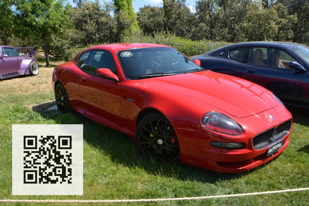 A red Maserati sports car parked on grass with a QR code in the foreground and another car in the background.