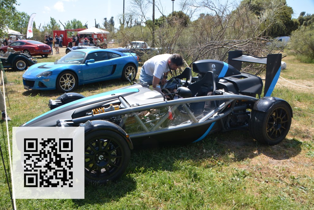 A person inspecting a black and blue lightweight sports car with an exposed chassis at an outdoor automotive event, with several other cars in the background.